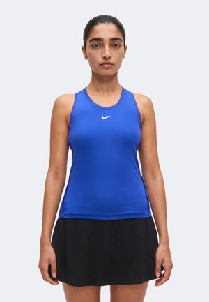 Kvinde står, iført en blå ærmeløs Nike sportstop og et sort skørt, vendt fremad mod en ensartet lys baggrund.