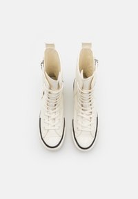 High-top sneakers in cream canvas with black rubber sole, front laces, side zipper, and textured interior. Rounded toe with toe cap.