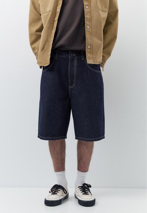 Mann trägt dunkelblaue Jeansshorts, eine braune Jacke über einem dunklen Hemd, weiße Socken und weiße Turnschuhe mit schwarzen Schnürsenkeln, steht vor einem schlichten Hintergrund.
