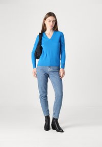Pulover mediu albastru cu decolteu în formă de V și mâneci lungi, asortat cu blugi deschiși la culoare și cizme negre cu toc. Modelul ține o geantă texturată neagră.