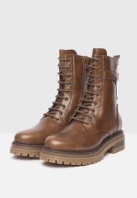 Une paire de bottes en cuir marron à lacets avec fermeture éclair sur le côté, sangle à boucle et semelles en caoutchouc épais sur fond blanc.