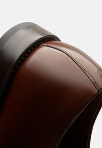 Scarpa in pelle marrone con design arrotondato. Presenta una superficie liscia, un tacco scuro a contrasto e cuciture visibili lungo la cucitura.