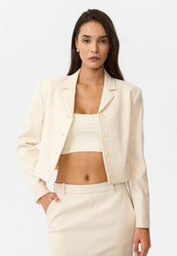 Blazer créme raccourci avec des revers, cinq boutons et poches. Porté sur un top ajusté assorti, présenté sur un fond neutre.