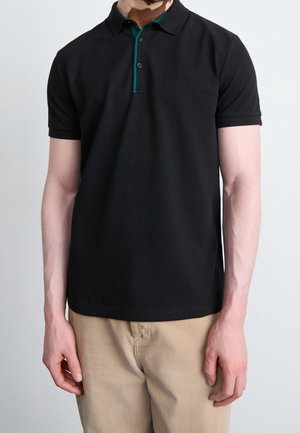 Musta poolopaita, jossa turkoosi kaulus ja kolmen napin läppä. Vasemmalla puolella näkyvä logo. Päällä vaaleat housut.