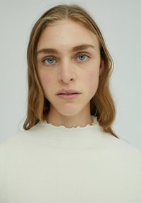 Helles beiges, geripptes Sweatshirt mit einem dezenten Rüschenausschnitt, kombiniert mit langen, welligen Haaren und klaren blauen Augen. Neutrale Hintergrund.