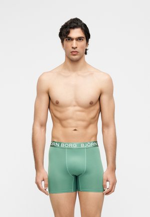 Man zonder hemd met kort, krullend donker haar die groene Björn Borg boxershorts draagt, staand tegen een effen lichte achtergrond.