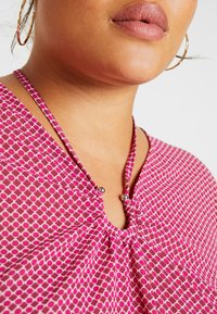 Haut rose à motif avec un décolleté en forme de goutte, doté d'un design froncé et d'accents en métal argenté près du col. Tissu à texture lisse.