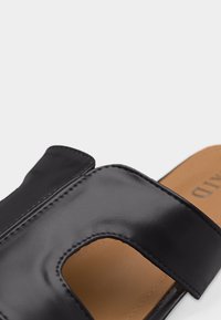 Sandale mule en cuir noir avec une large bride présentant une grande découpe rectangulaire, une semelle intérieure rembourrée beige et des détails de couture visibles.