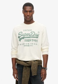 Crème lange mouw shirt met groene bedrukte tekst en logo. Donkere jeans met een bruine gebreide trui om de taille geknoopt. Zwarte horloge om de pols.