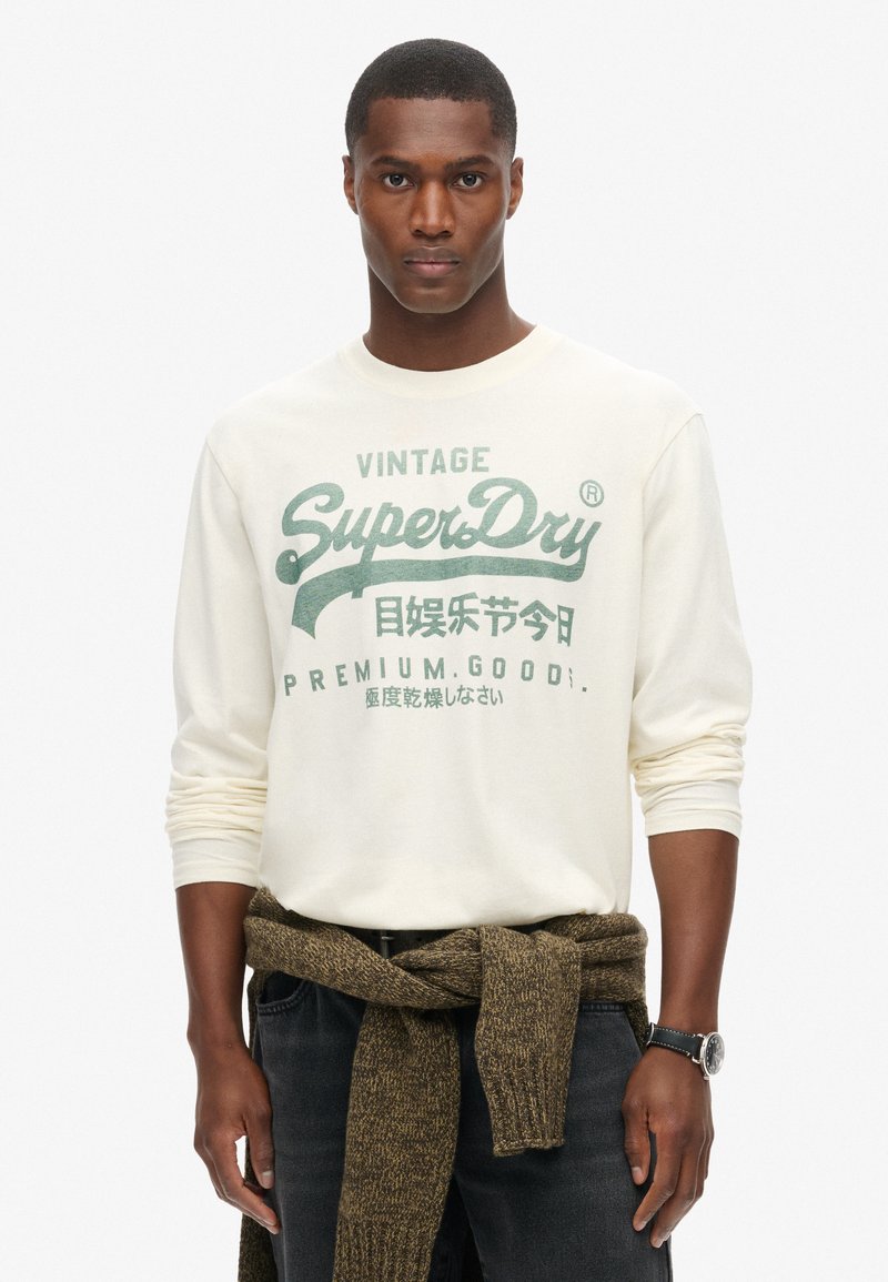 Crème lange mouw shirt met groene bedrukte tekst en logo. Donkere jeans met een bruine gebreide trui om de taille geknoopt. Zwarte horloge om de pols.