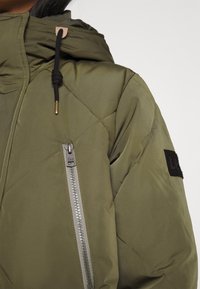 Gros plan d'une personne portant une veste matelassée vert olive avec capuche, cordon noir, poche zippée argentée en diagonale et écusson noir sur la manche.