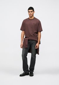 Camiseta corta de manga en color marrón con cuello redondo, combinada con jeans oscuros. Zapatos negros tipo slip-on complementan el atuendo, creando un look casual.