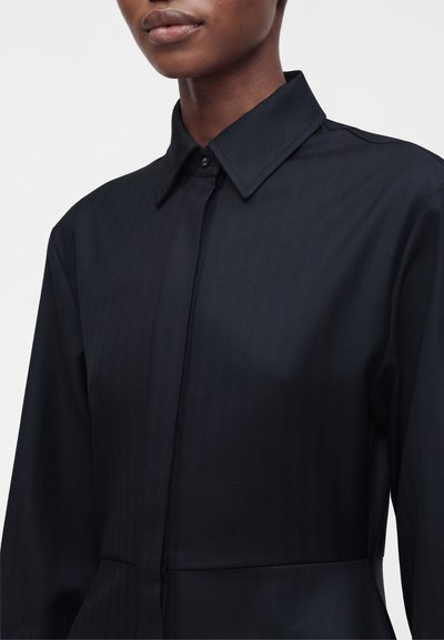 Personne portant une chemise bleu marine foncé à col boutonné au niveau du cou, vue des épaules au milieu du torse sur un fond uni.