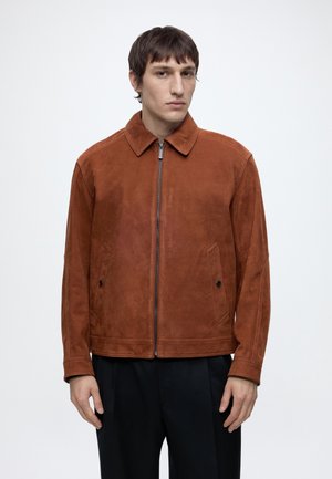 Mann trägt eine braune Wildlederjacke mit Reißverschluss und Kragen sowie schwarze Hose, steht vor einem schlichten weißen Hintergrund.