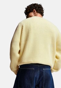 Geel geribbelde gebreide trui met een ontspannen pasvorm en verlaagde schouders, gecombineerd met donkerblauwe denim jeans. De textuur is zacht en chunky.