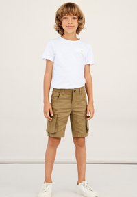 T-shirt blanc à manches courtes avec un détail de poche, associé à un short cargo kaki avec poches latérales et fermeture à bouton. Baskets blanches.