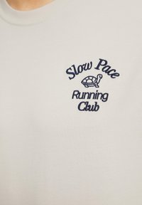 Lichtgrijze katoenen T-shirt met marineblauwe geborduurde tekst: "Slow Pace Running Club" met een schildpadgraphic. Klassiek ronde hals ontwerp.