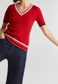 Top corto de punto acanalado rojo con cuello en V, que presenta acentos de rayas blancas y azul marino en el cuello y el dobladillo. Combinado con denim oscuro.