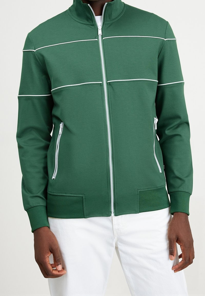 Giacca verde con zip e dettagli a strisce bianche. Tessuto liscio, colletto alto e tasche laterali con zip. Design aderente, polsini e orlo a coste.