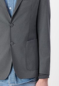 Grijze blazer van gestructureerde stof, met twee knopen, een borstzak en een relaxte pasvorm over een denim shirt.