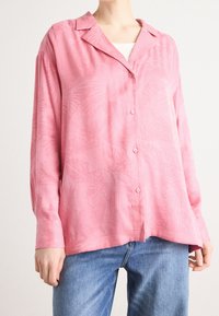 Personne portant une chemise ample rose à boutons avec des motifs subtils de plumes et un jean bleu clair sur un fond uni.