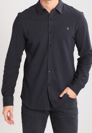 Mann trägt ein enganliegendes, langärmliges schwarzes Hemd mit Knöpfen und kleinem orangem Logo auf der linken Brust sowie schwarze Hose, mit der Hand in der Hosentasche.