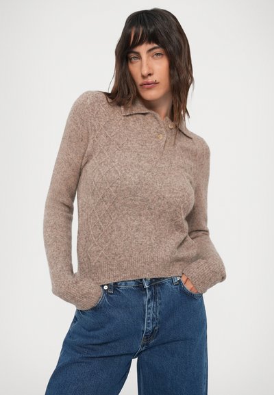 Mujer con cabello oscuro, vestida con un su�éter de punto beige con cuello y jeans azules, con las manos en los bolsillos, frente a un fondo claro y liso.