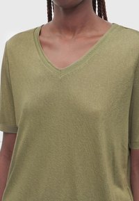 Person trägt ein olivgrünes Kurzarmshirt mit V-Ausschnitt, geflochtene Haare sichtbar, beschnitten unterhalb des Mundes und oberhalb der Taille.
