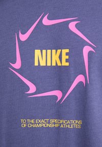 Violet medvilnės audinys turi rožinius Nike logotipus, išdėstytus apskritimu, su žodžiu "NIKE" geltonais rašmenimis ir papildomu tekstu žemiau.