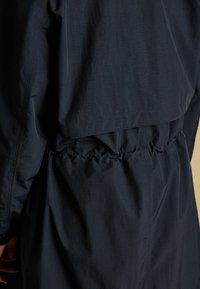 Jachetă ușoară de culoare albastru marin, cu o talie strânsă, o textură netedă și un design stratificat, având detalii elastice la margine.