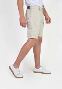Shorts en coton beige avec des revers, poches avant et une poche arrière à rabat, associés à des baskets blanches avec une semelle marron.
