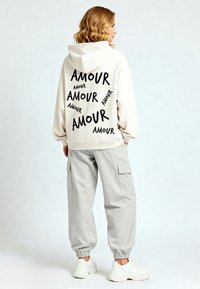 Felpa beige chiaro con motivo testuale "AMOUR" nero, abbinata a pantaloni cargo grigi chiari e sneakers bianche. Tessuto morbido e vestibilità rilassata.