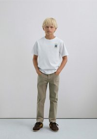 Blonde jongen die staat met zijn handen in zijn zakken, gekleed in een wit T-shirt met grafische druk, beige broek en bruine sneakers tegen een effen lichte muur.