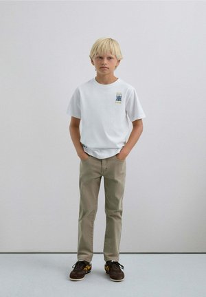 Niño rubio de pie con las manos en los bolsillos, vistiendo una camiseta blanca con un gráfico, pantalones beige y zapatillas marrones contra una pared clara.