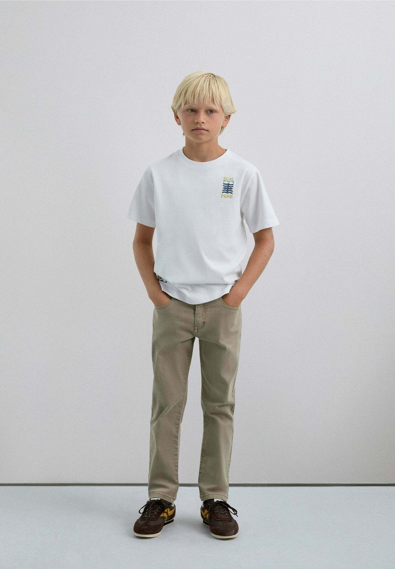 Blonde jongen die staat met zijn handen in zijn zakken, gekleed in een wit T-shirt met grafische druk, beige broek en bruine sneakers tegen een effen lichte muur.