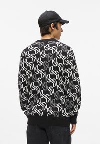 Melnbalts adījuma džemperis ar atkārtotu logotipa rakstu, ribinātas aproces un apakšmalu, valkāts ar melnu tīkla cepuri, redzams no aizmugures.