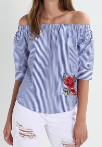 Blusa off-shoulder a righe blu e bianche con maniche corte, caratterizzata da ricami floreali rossi sul lato sinistro e una vestibilità rilassata.