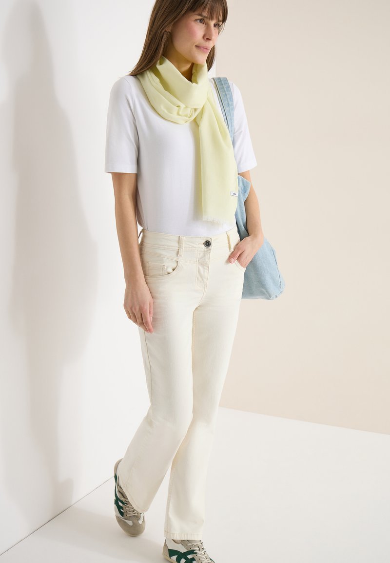 Femme debout les mains dans les poches, portant un t-shirt blanc, une écharpe jaune pâle, un pantalon blanc cassé, des baskets et un sac à bandoulière bleu clair.