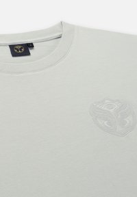 Lichtgrijs t-shirt van zacht stof, met een ronde halslijn en een reliëflogo op de linkerkant van de borst. Gladde textuur, eenvoudig ontwerp.