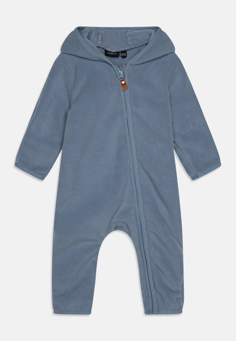 Lyseblå fleece overalls med hette, med lange ermer, glidelås foran og brun skinnaccent. Myk, teksturert stoff.