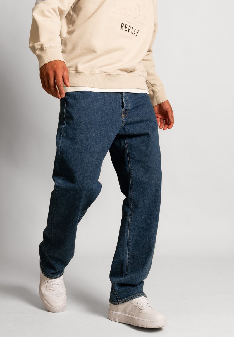 Jeans in denim blu con vestibilità ampia, dotati di una zip standard e cuciture a contrasto, abbinati a sneakers bianco sporco e a una felpa beige chiaro.