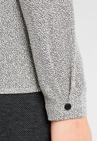 Long-sleeve blouse in a beige fabric with a black speckled pattern, featuring a button closure at the cuff. Textured, lightweight material.