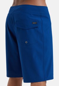 Blå badbyxor med bakficka, platt sömnad och en liten logotagg. Materialet verkar lätt och smidigt.