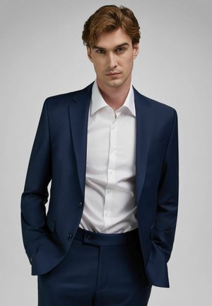 Junger Mann mit hellbraunen Haaren, der einen marineblauen Anzug und ein weißes Hemd trägt, die Hände in den Taschen, schaut nach vorne vor einem schlichten Hintergrund.