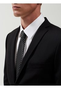 Chaqueta de traje negra con solapa de muesca, llevada sobre una camisa blanca. La corbata negra presenta rayas blancas. Detalle del pañuelo de bolsillo en la parte izquierda.