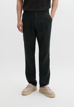 Mężczyzna w czarnych spodniach o prostych nogawkach, beżowych casualowych butach sznurowanych, stoi z jedną ręką w kieszeni na białym tle.