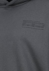 Dark gray fabric with small black text reading "Happiness matters for all of us!... 24H // SELF CARE // EVERY DAY" near the neckline.