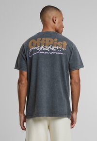 Šedé bavlnené tričko s okrúhlym výstrihom, na zadnej strane s potlačou horskej grafiky a textom "OffPist" v béžovej a purpurovej farbe.