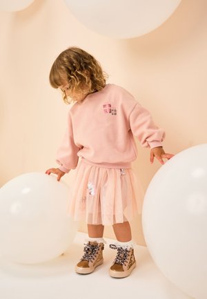 Sweat-shirt rose avec encolure arrondie, associé à une jupe transparente à plusieurs couches avec des détails pastels. Bottes à imprimé léopard avec des lacets décoratifs.