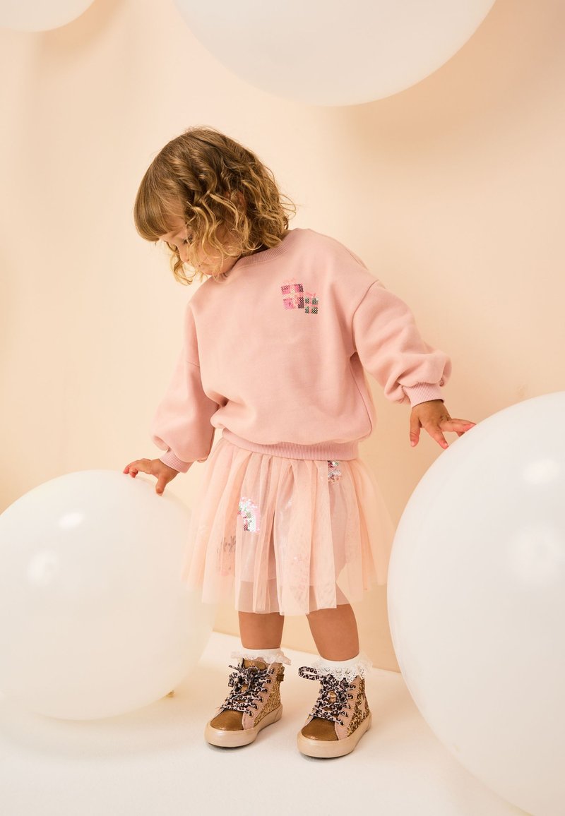 Sweat-shirt rose avec encolure arrondie, associé à une jupe transparente à plusieurs couches avec des détails pastels. Bottes à imprimé léopard avec des lacets décoratifs.